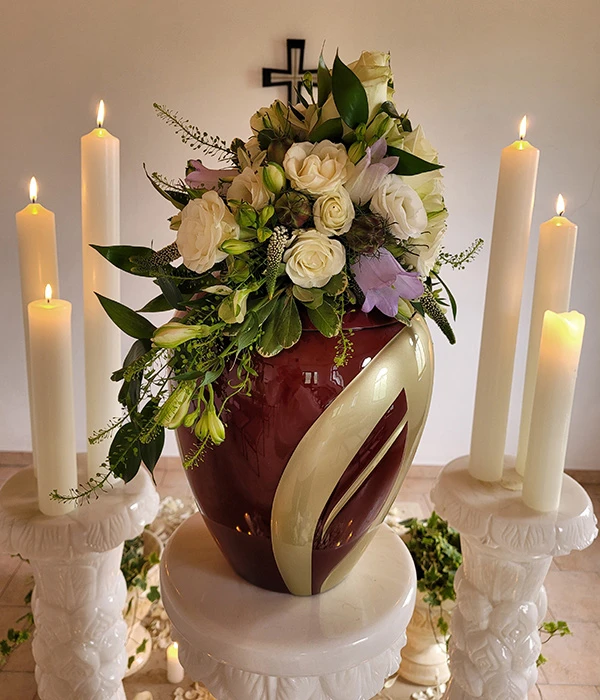 Weinrote goldverzierte Urne mit Blumenschmuck auf Marmorsäulen und Kerzen – Remuna Bestattungen Halle (Saale)